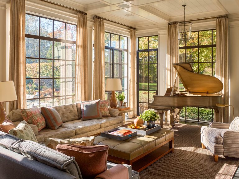 Sunlit living room with large windows, beige sofas with colorful cushions, a wooden coffee table with books and a potted plant, two lamps, a striped armchair, and a grand piano in the corner; trees are visible outside.