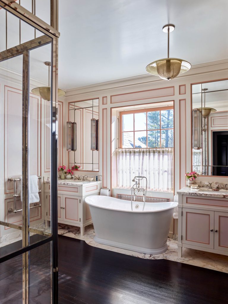 Elegant bathroom with dark wood floor, white freestanding tub, and gold fixtures. Soft pink and white paneled walls, large window with sheer curtain, double vanities, tall mirrors, and small floral arrangements create a bright, classic look.