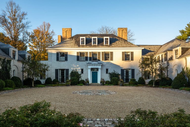 A large, elegant white mansion with symmetrical architecture, black shutters, and a teal front door sits behind a circular gravel driveway. The house has two prominent brick chimneys and is surrounded by manicured greenery and autumn trees.