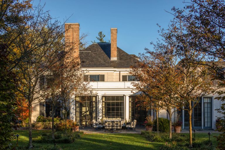 A large, elegant house with two tall brick chimneys, white walls, and tall windows sits among trees with autumn foliage. Patio furniture is arranged on a stone terrace, and there is manicured greenery in the foreground under a clear blue sky.