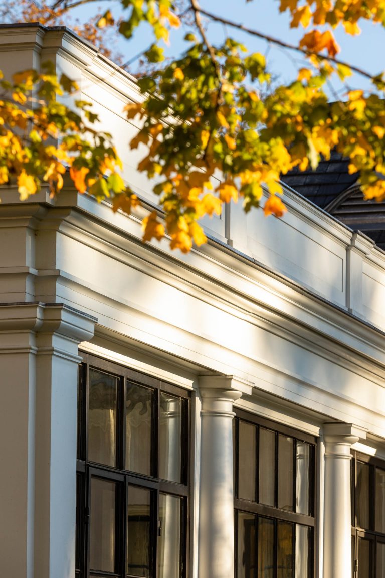 A close-up of a classical building facade with white columns and large black-framed windows, partially shaded by tree branches with green and yellow autumn leaves in sunlight.
