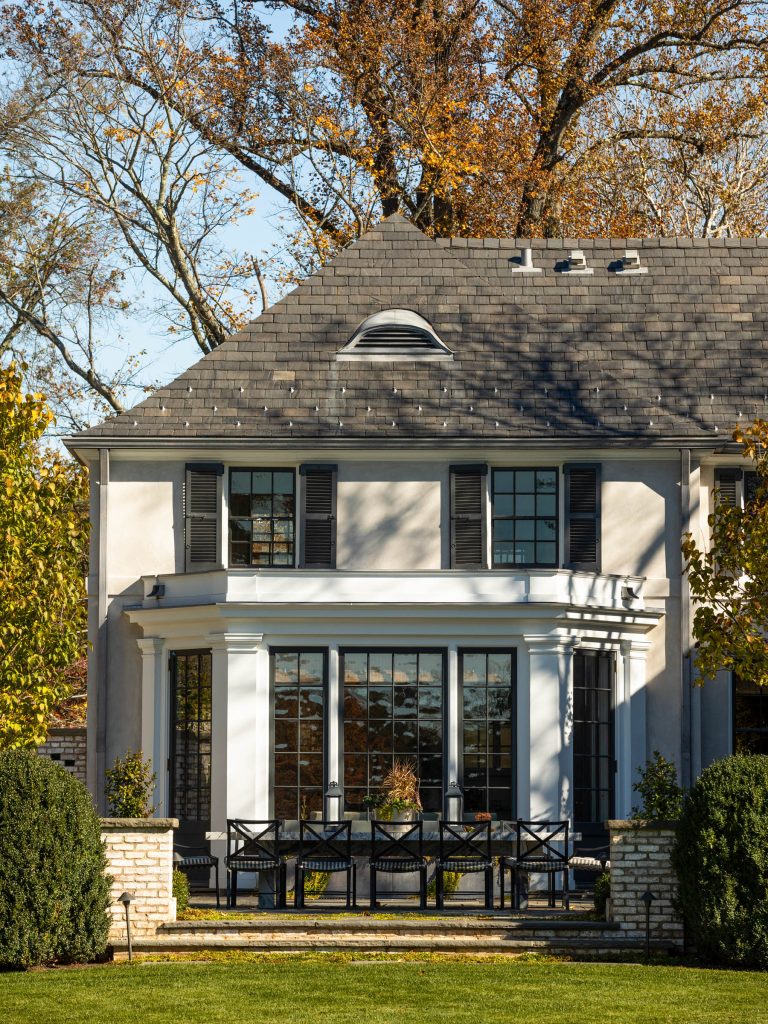A two-story house with gray shingles, white walls, black window shutters, and a prominent bay window. Large black-framed glass doors open to a patio with black chairs and potted plants. Manicured lawn and trees surround the home, with autumn foliage in the background.