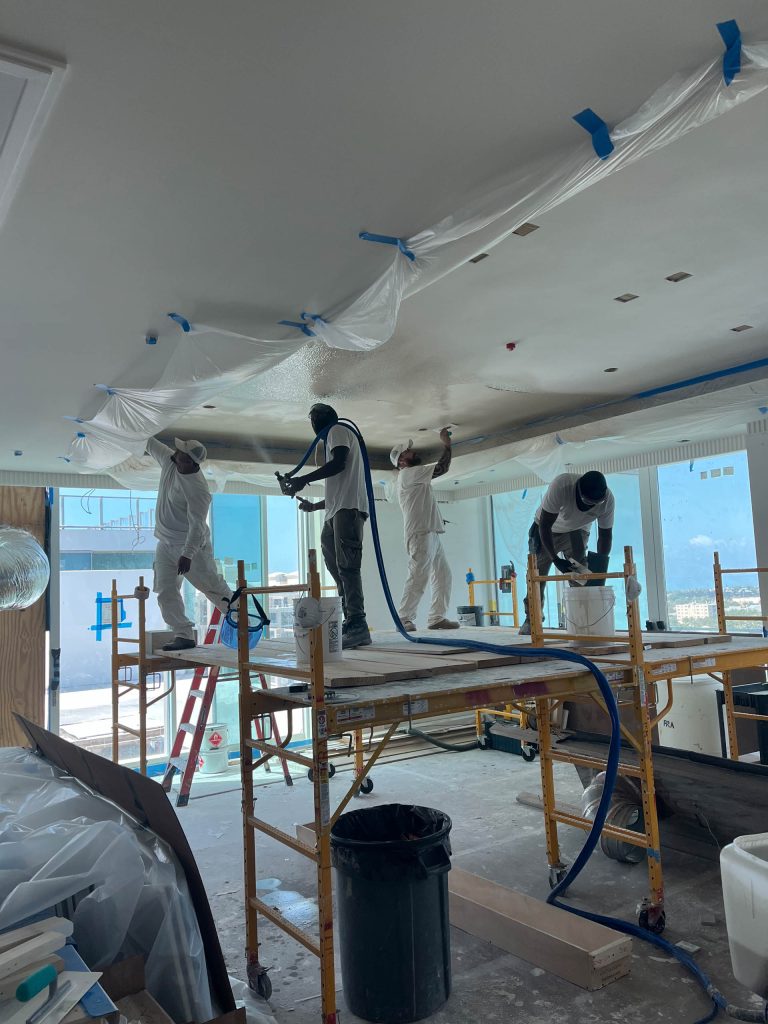Four workers in white protective clothing stand on scaffolding, spray painting or plastering the ceiling of a Miami Beach penthouse. Plastic covers parts of the area, ladders and tools are visible, and a large window shows a blue building outside.