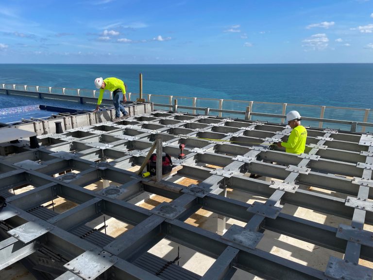 Two construction workers in safety gear work on a steel framework atop a Miami Beach penthouse. One kneels on the grid while the other stands, both in white helmets and neon yellow shirts. Blue sky and a bridge over water are visible in the background.