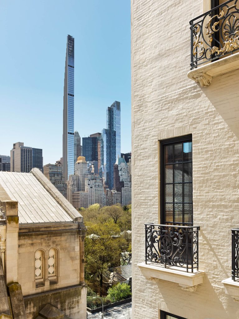 View of New York’s cityscape with tall modern skyscrapers in the background and older, ornate buildings—some undergoing Fifth Avenue Renovation—with wrought-iron balconies and decorative windows in the foreground. Trees and a clear blue sky complete the scene.
