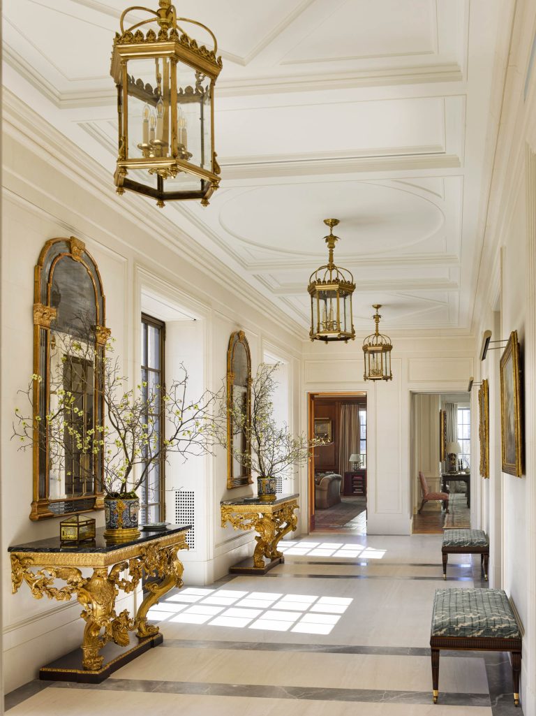 Elegant hallway in this Fifth Avenue Renovation features cream walls, a high coffered ceiling, and large windows. Ornate gold mirrors and consoles with vases of branches line the wall, reflecting New York sophistication throughout.