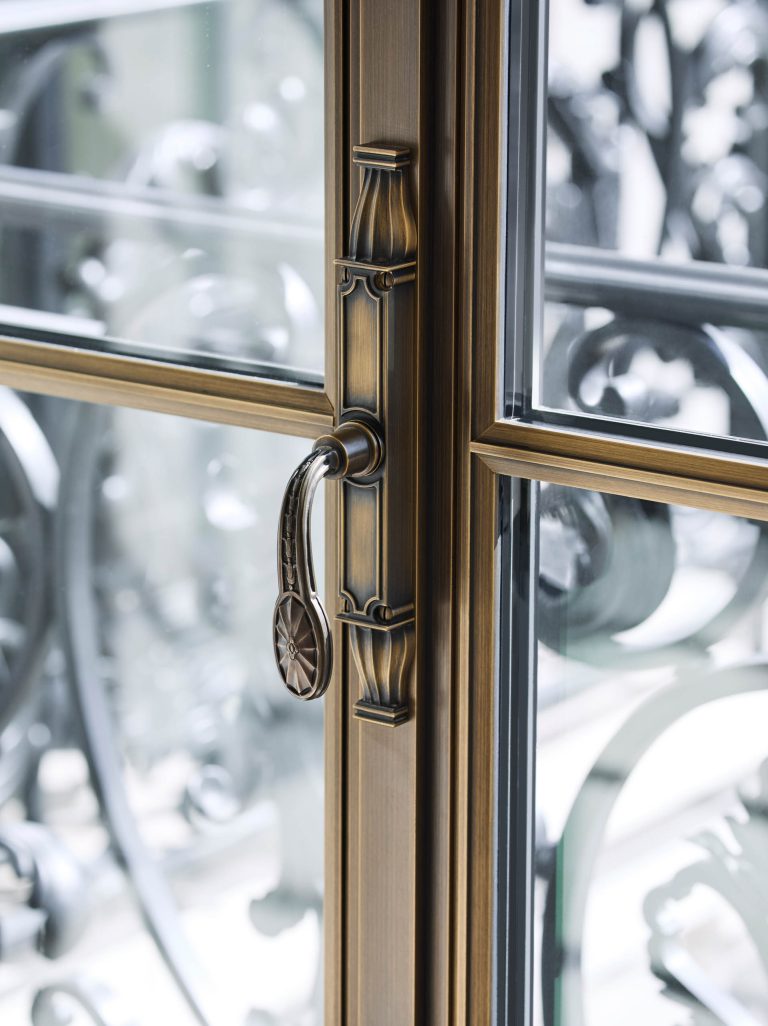 Close-up of an ornate, antique brass door handle and latch on a glass door, featuring intricate details. Through the clear glass, an elegant wrought iron balcony railing appears—part of a Fifth Avenue Renovation in New York.
