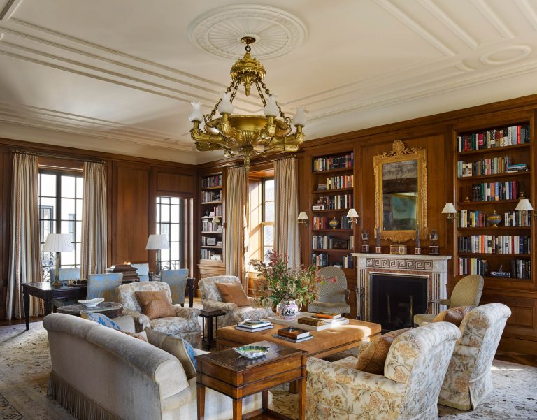 Elegant living room in a sophisticated Fifth Avenue Renovation, with wood-paneled walls, ornate ceiling, and a grand gold chandelier. Plush armchairs, sofas, and tall windows evoke classic New York charm in this inviting space.