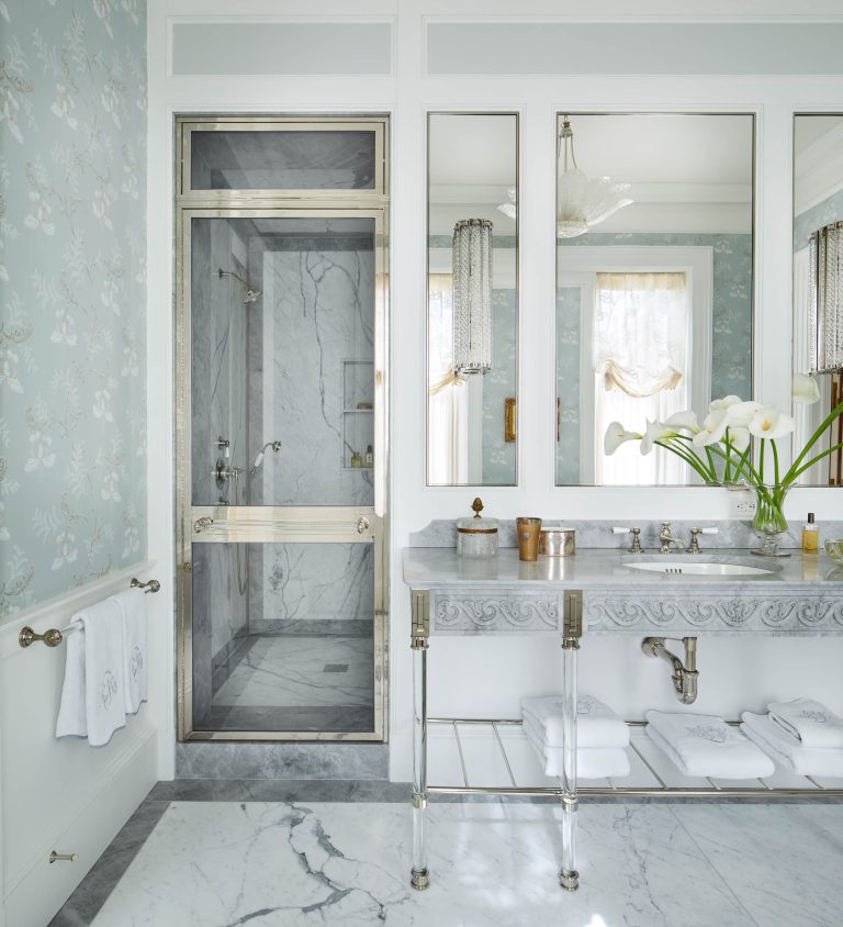 Elegant bathroom in a Fifth Avenue Renovation with a marble shower and gold trim, double sinks with a gray marble counter, large mirrors, and white calla lilies. Light blue floral wallpaper and natural light give this New York space a soft, luxurious touch.