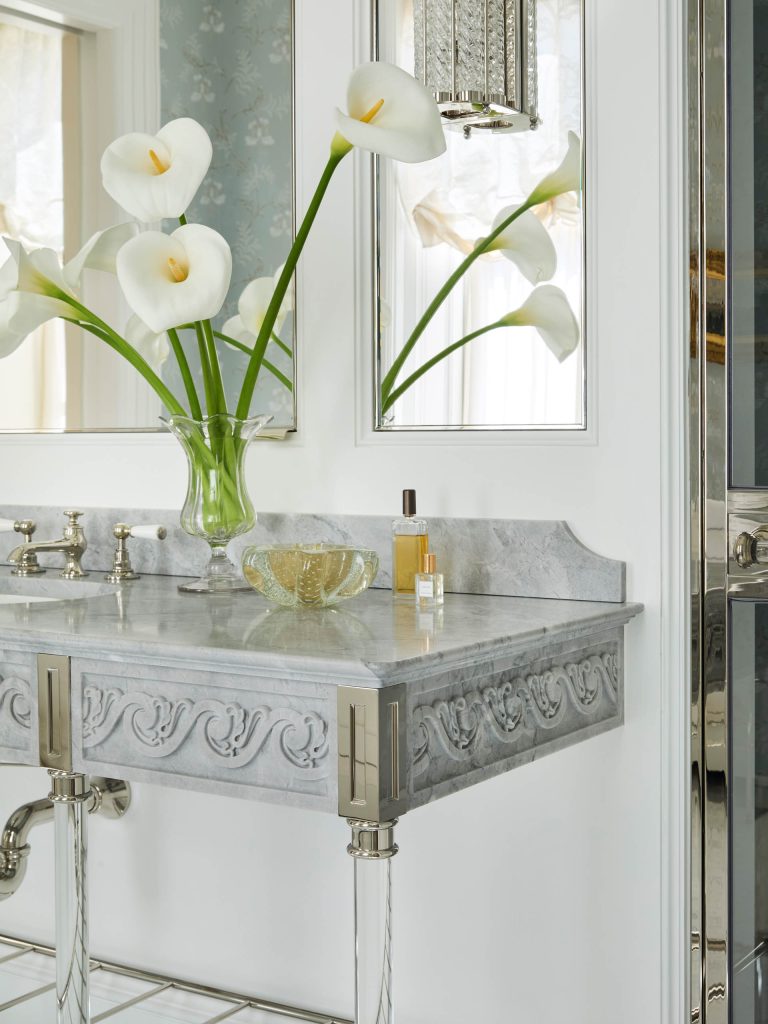 A marble bathroom vanity with ornate detailing, part of a Fifth Avenue Renovation, holds a vase of white calla lilies, a perfume bottle, and a glass dish. A mirror and metal fixtures reflect light; sheer curtains frame a window in the background.
