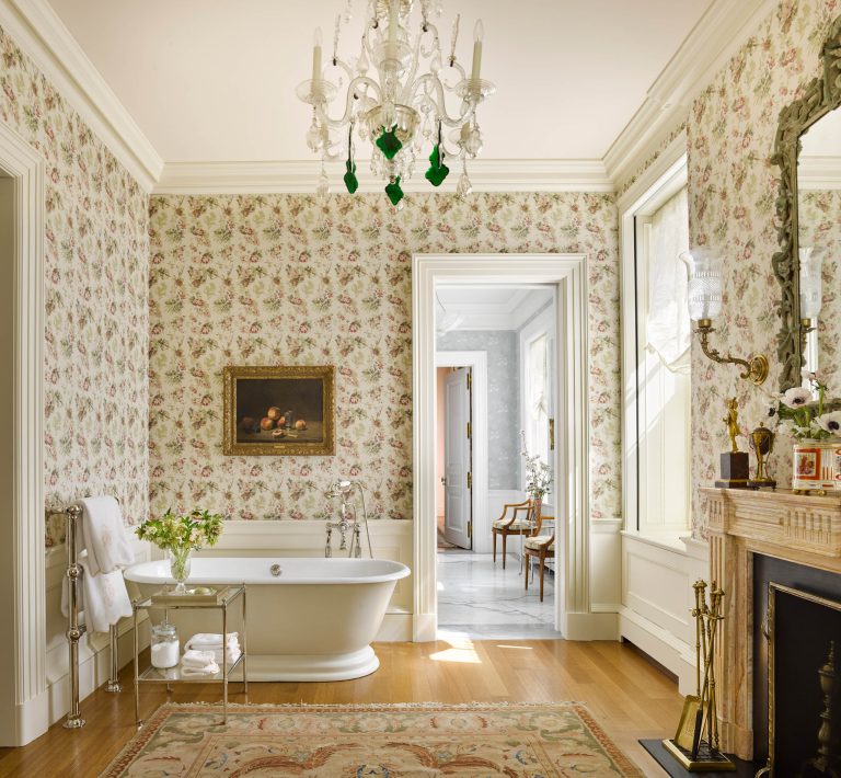 Elegant vintage bathroom with floral wallpaper, freestanding white tub, ornate chandelier, and a fireplace with a large mirror—capturing the charm of a Fifth Avenue Renovation in New York. An open door reveals a bright adjoining room with chairs and marble floor.