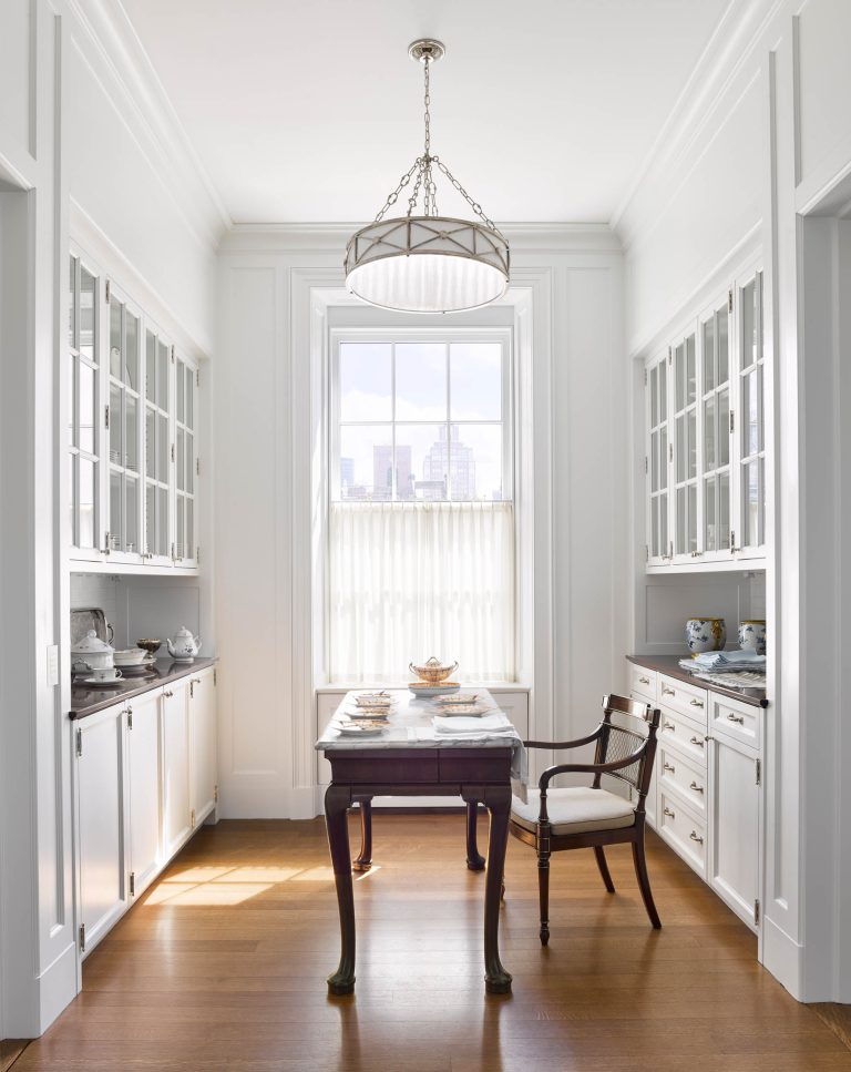 A bright, elegant butler’s pantry with glass-front cabinets, white walls, and wood floors sits at the heart of this Fifth Avenue Renovation. Sunlight streams through a large window with New York city views, highlighting china on the shelves.