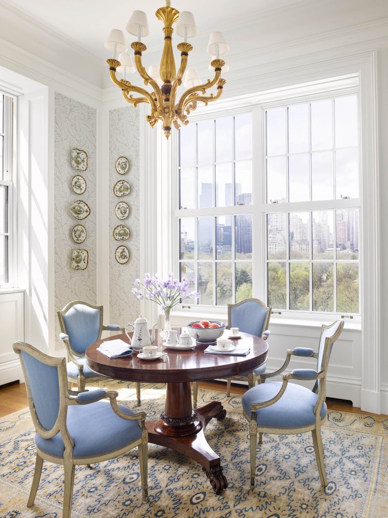 A bright dining room with large windows overlooking a park sets the stage for this Fifth Avenue Renovation. A round wooden table, blue-upholstered chairs, gold chandelier, and patterned rug offer classic New York elegance and charm.