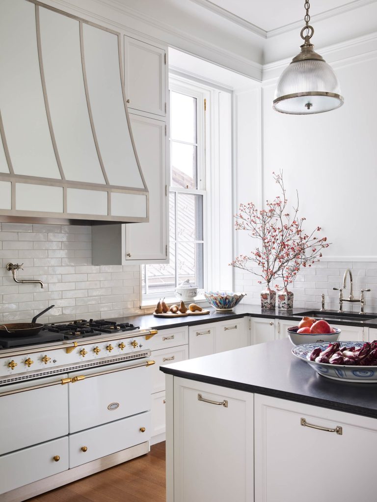 A bright, elegant kitchen inspired by a Fifth Avenue Renovation features white cabinets and gold hardware, a vintage-style oven, and a large range hood. Subway tiles and tall windows fill the New York space with abundant natural light.