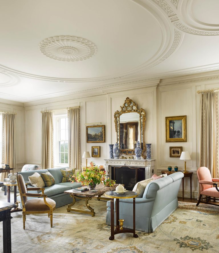 A grand, elegant living room inspired by a Fifth Avenue Renovation in New York, with ornate ceiling molding, large windows draped in beige curtains, pale blue sofas, a marble fireplace, antique tables, classic paintings, and fresh flowers on the coffee table.