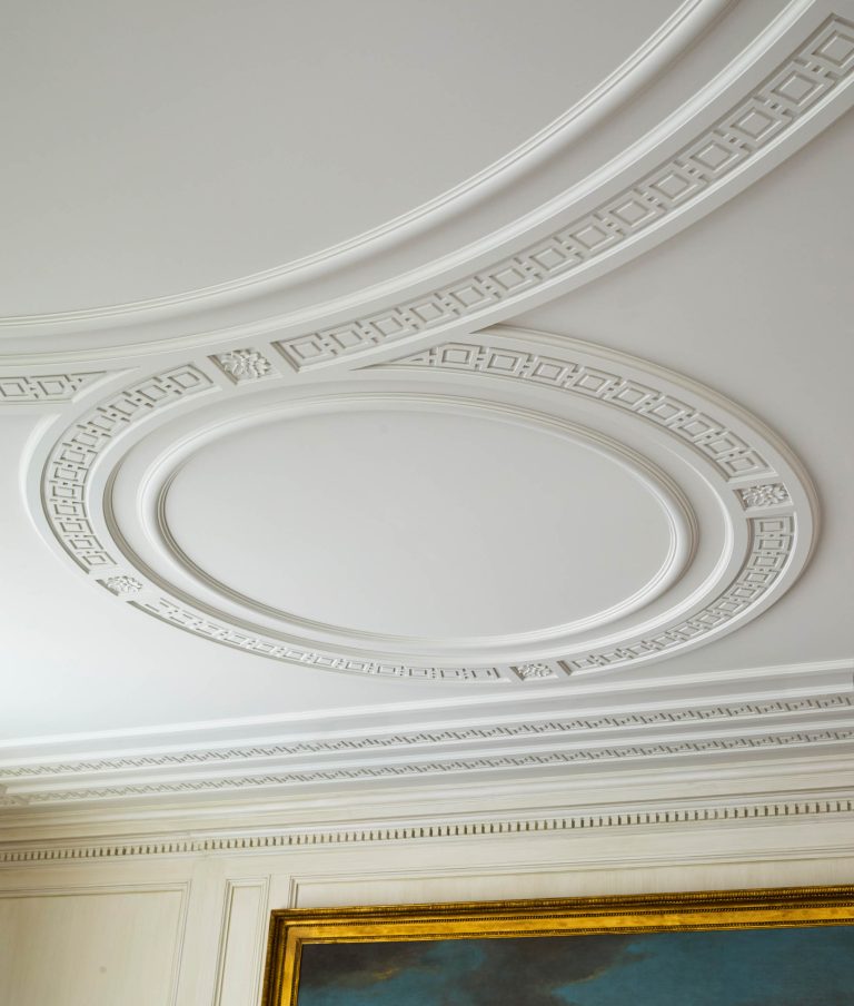 Elegant white ceiling with intricate decorative molding featuring concentric oval and circular patterns with Greek key motifs—a refined touch to this Fifth Avenue Renovation. Cream-colored paneled walls and a gold-framed painting complete the New York sophistication.