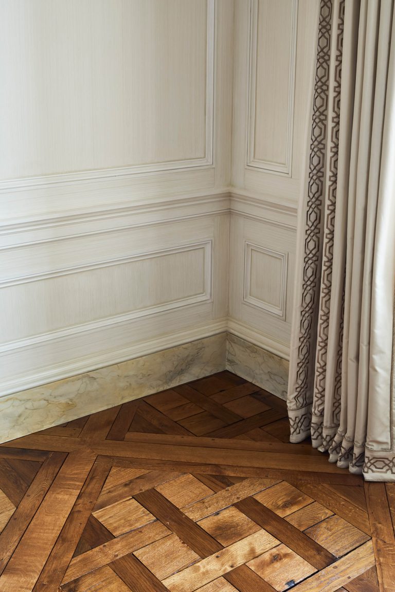 Corner of a room from a Fifth Avenue Renovation in New York, featuring cream paneled walls, ornate dark wood parquet flooring, a marble baseboard, and heavy beige curtains with an intricate patterned edge.