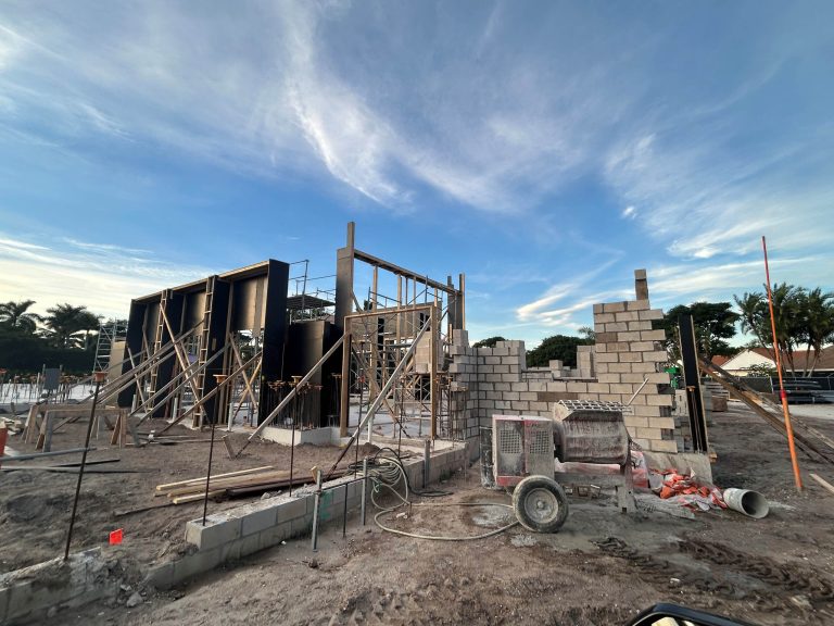 A construction site on a Wellington Equestrian Estate with partially built concrete block walls and metal supports. Scaffolding surrounds unfinished structures while construction equipment and materials are scattered across the dirt under a blue sky. Trees line the background.