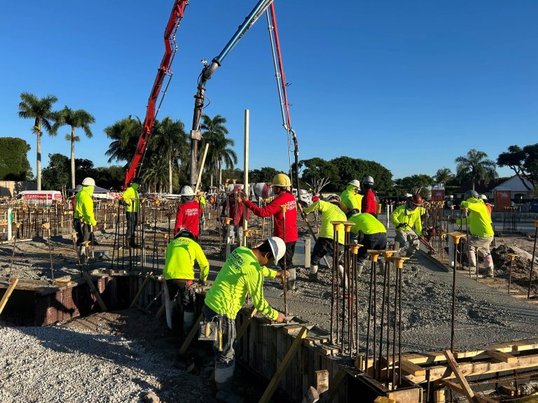 Construction workers in bright safety gear pour concrete into a large foundation at a Wellington equestrian estate. The busy site features red concrete pumping machines, steel rods, and wooden forms beneath palm trees and a clear blue sky.