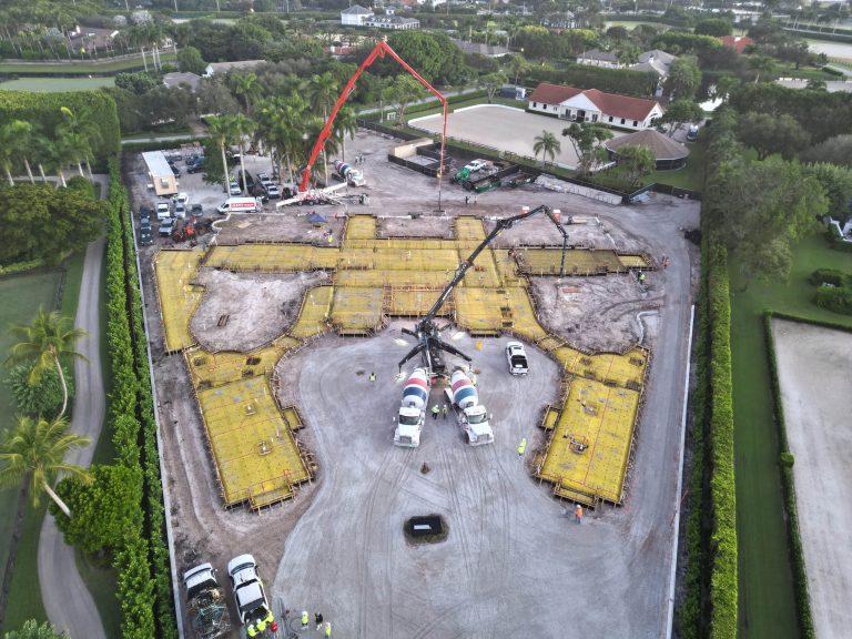 Aerial view of a large Wellington construction site with yellow forms and rebar outlining a building foundation. Concrete trucks, cranes, and workers are present. The site is surrounded by trees, grass, and nearby equestrian estate buildings.