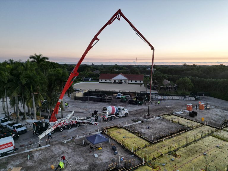 Aerial view of a construction site at sunset in Wellington, with a large red concrete pump truck pouring concrete for an Equestrian Estate foundation. Workers in safety gear, equipment, vehicles, palm trees, and a white building are visible.