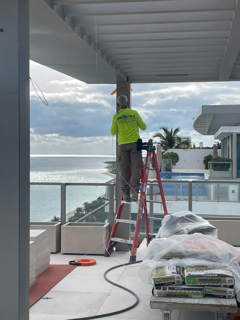 A worker in a neon green shirt and hard hat stands on a red ladder, working on a beam of a modern penthouse balcony overlooking Miami Beach. Construction materials lie scattered, framed by tall glass railings and contemporary buildings nearby.