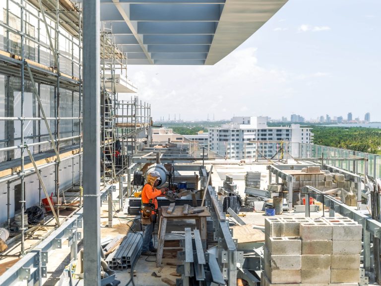 A construction worker in an orange shirt and hard hat works at a table on a high-rise penthouse under construction, surrounded by scaffolding and beams, with Miami Beach’s cityscape and trees visible in the background beneath a clear sky.