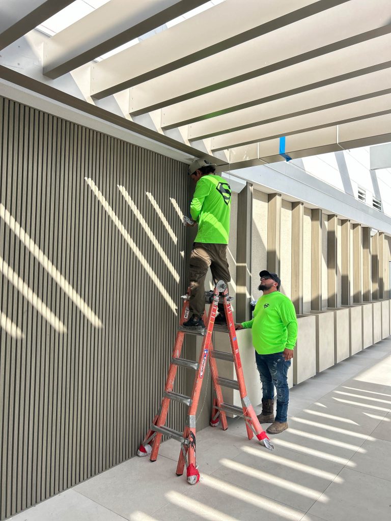 Two construction workers in neon green shirts work indoors on a Miami Beach penthouse; one stands on a ladder adjusting a wall fixture while the other watches. Sunlight casts striking striped shadows of structural beams onto the wall and floor.