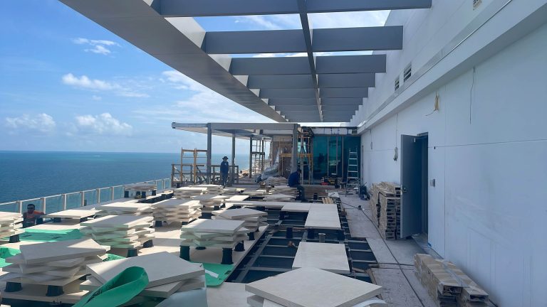 A penthouse rooftop terrace under construction overlooks the ocean in Miami Beach. Stacks of tiles and building materials cover the floor, while workers and ladders are visible near the glass railings beneath a partial canopy and partly cloudy sky.