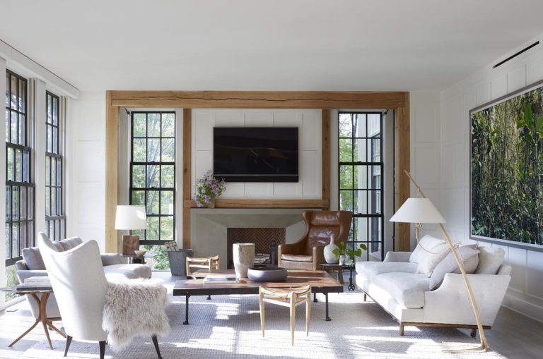 Bright modern shingle-style living room with large windows, white walls, and a fireplace with a wooden mantel. Princeton-inspired décor includes tan sofas, leather armchair, wooden coffee table, wall-mounted TV, and a striking nature photo on the right wall.
