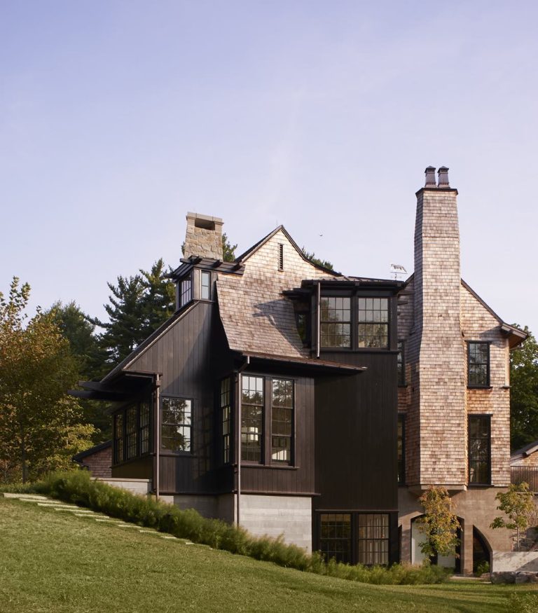 This three-story Princeton home showcases Modern Shingle style with a steep shingle roof, tall stone chimneys, and black-framed windows. Dark wood siding and shingles blend beautifully into the grassy slope and tree-lined backdrop under a clear sky.
