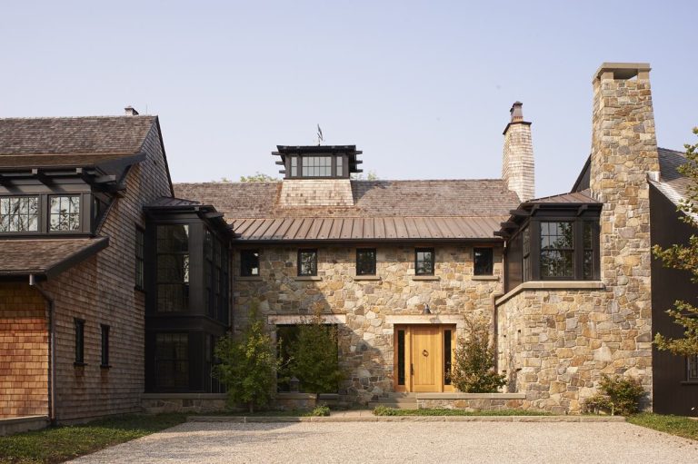 A large house with a modern shingle roof and stone exterior walls sits in Princeton, featuring a wooden front door, black-trimmed windows, and a tall stone chimney. Small trees and shrubs line the gravel driveway leading to the entrance.