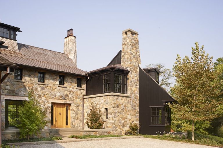 A large Princeton home with stone walls, a wooden front door, and a tall stone chimney. Black-trimmed windows and Modern Shingle siding accent one section. Trees and shrubs frame the entrance, with a gravel driveway under a clear sky.