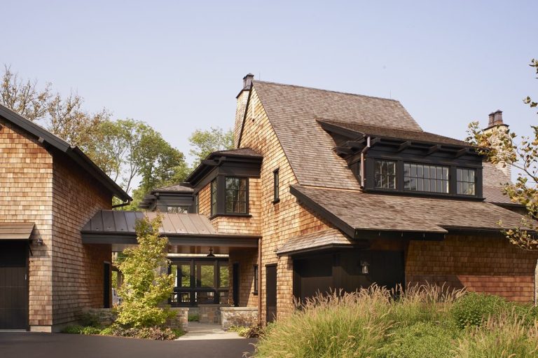 A modern Princeton house with Modern Shingle siding and a steep roof, connected by a glass entryway. Large windows are framed in black. Tall grasses and a small tree grow in front, under a clear blue sky.