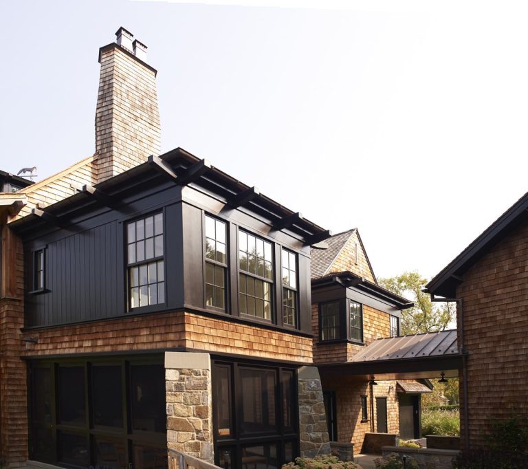 A two-story Modern Shingle house in Princeton with stone on the lower level, cedar shingles, and black trim framing expansive windows. A glass walkway connects two sections, while a tall stone chimney and abundant natural light enhance the exterior.