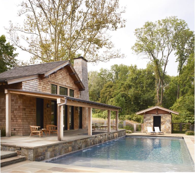 A Modern Shingle wooden house with a stone patio sits beside a rectangular swimming pool. Two wooden chairs are on the patio, and a lounge chair is next to the pool. Lush green trees surround the Princeton retreat, with a stone pool house in the background.