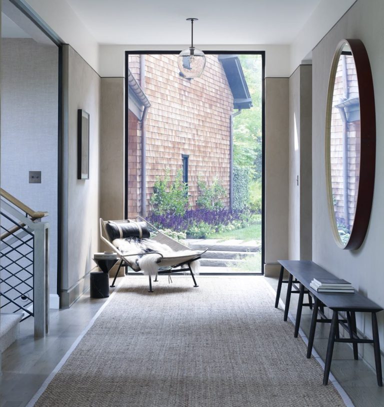 A modern hallway with a large glass door frames a Modern Shingle house and greenery. Inside, a black lounge chair with fur throw, bench with books, round mirror, light walls, and woven rug evoke an inviting Princeton elegance.