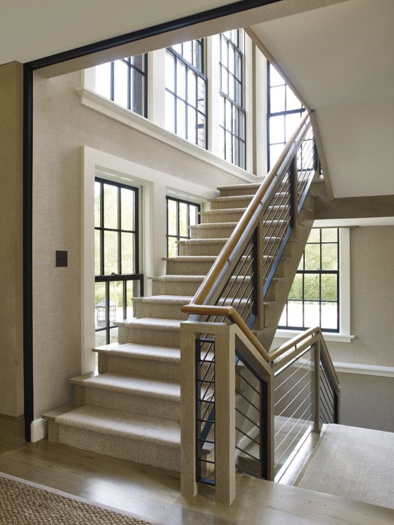 A bright, modern staircase with light carpeted steps, wooden handrails, and black metal balusters. Large, black-framed windows evoke a Modern Shingle style, flooding the Princeton home’s neutral walls and hardwood floor with natural light.