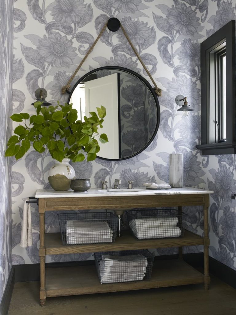 A Modern Shingle bathroom with floral wallpaper, a round mirror hanging by a rope, wooden vanity and white sink, leafy plant, open shelves with wire baskets of rolled towels, and a black-framed window on the right wall.