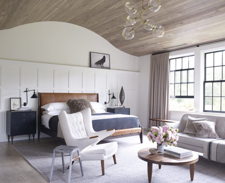 A modern shingle bedroom in Princeton featuring a curved wooden ceiling, bed with leather headboard, dark blue nightstands, white armchair and ottoman, light-gray sofa, round wooden coffee table, large windows with beige curtains, and wall art above the bed.