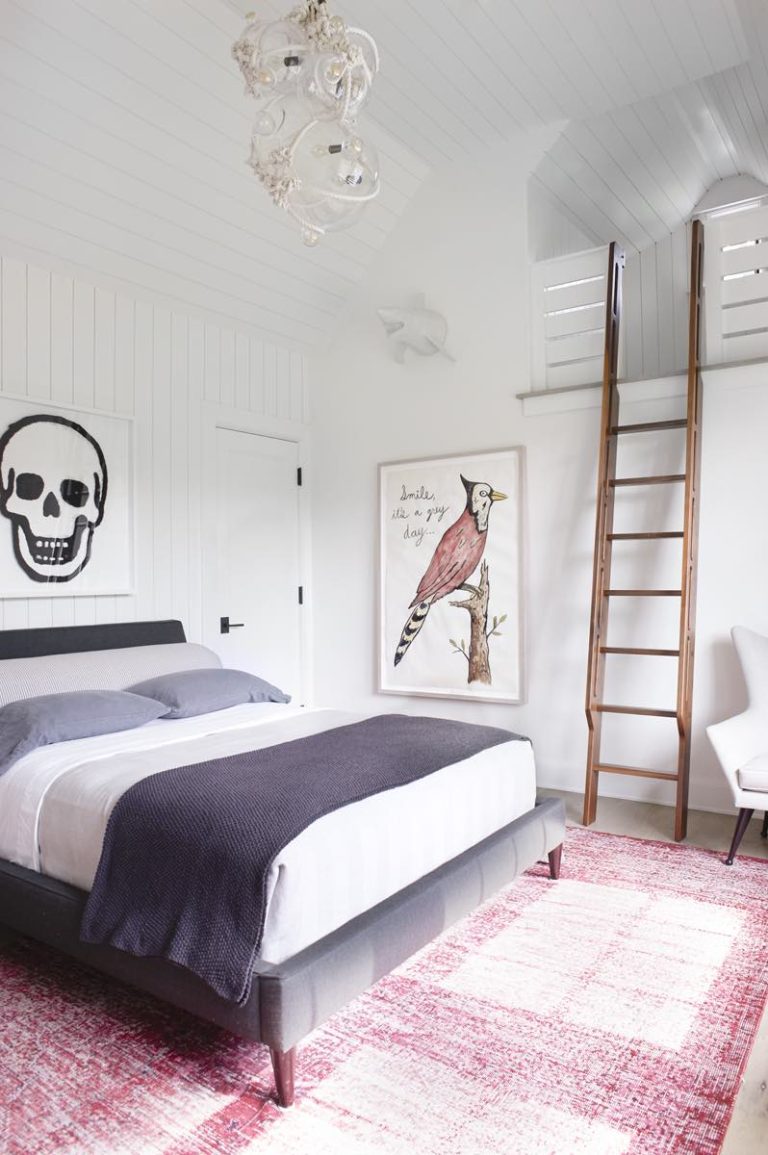 A bright Princeton bedroom with white paneled walls, a dark bed with gray and white bedding, a red patterned rug, a wooden ladder to a loft, skull and bird wall art, a white chair, and a modern glass light fixture in Modern Shingle style.