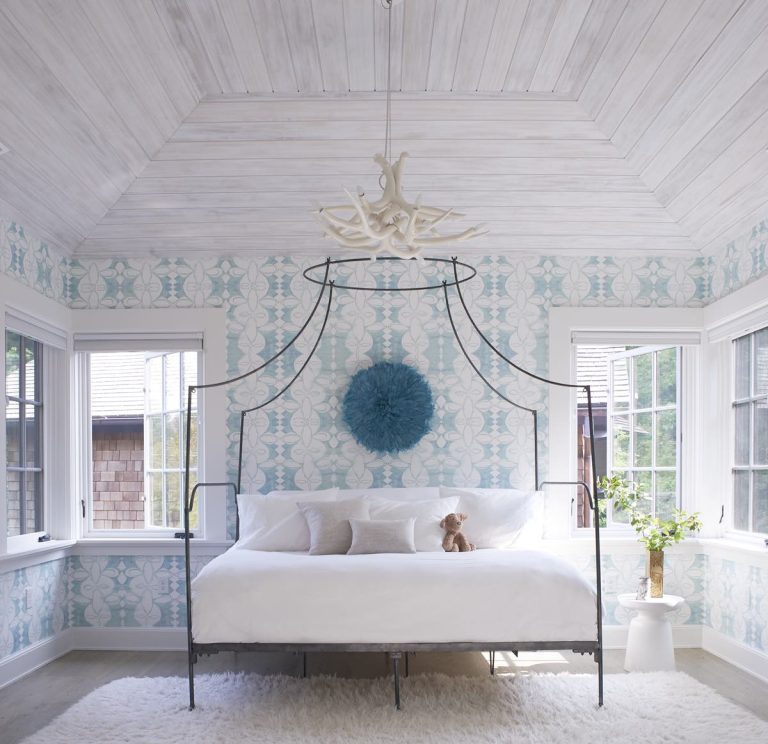 Bright bedroom with Modern Shingle-inspired blue-and-white wallpaper, a canopy bed with white bedding, and light wood-paneled ceiling. Blue feathered wall art hangs above the bed; a plush toy and vase of flowers add Princeton charm by the window.