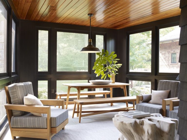 A sunroom with large black-framed windows and a wood ceiling evokes Modern Shingle style. Two wooden armchairs with gray cushions, a wooden bench and table, and lush greenery outside create a serene Princeton-inspired retreat.