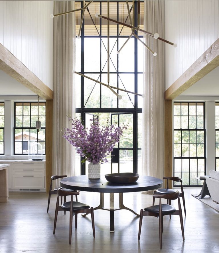 Bright, modern dining space with tall windows and sheer curtains in a stylish Modern Shingle home. A round black table with four chairs surrounds a large vase of pink flowers, while a gold chandelier hangs from the high ceiling. Light floods the room.