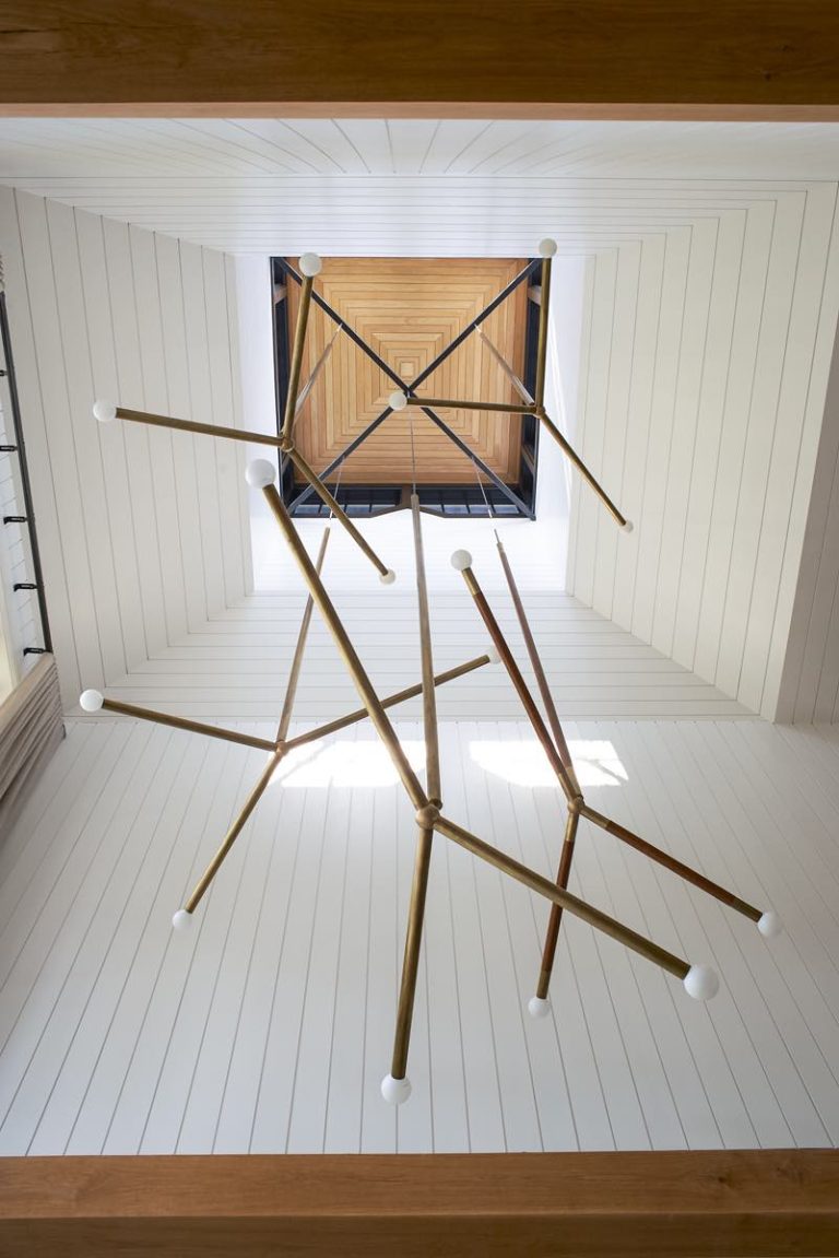 View looking up at a modern chandelier with intersecting gold rods and white globes, hanging in a tall, white-paneled shaft. The ceiling above has a Princeton Modern Shingle style square wooden inset that creates a geometric visual effect. Natural light highlights the scene.