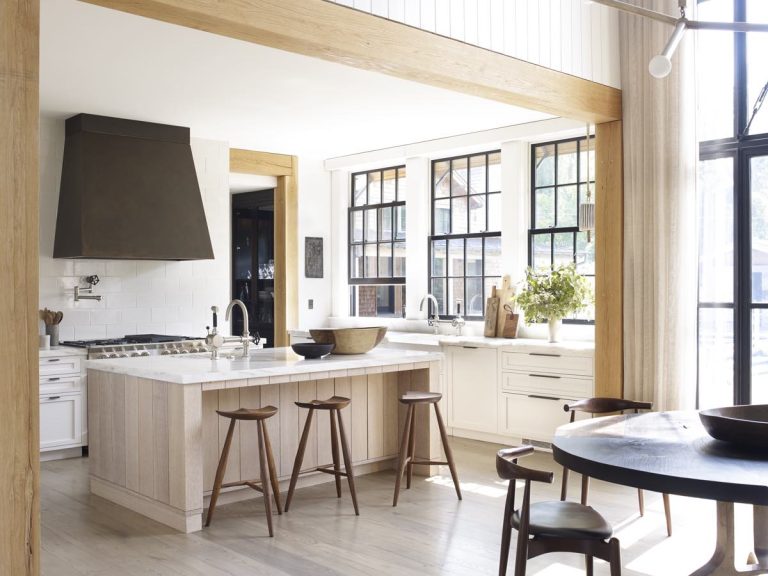Bright, modern Princeton kitchen with large windows, a central island with wooden stools, white cabinetry, marble countertops, stainless steel appliances, a dark metal range hood, light wood accents, round dining table, and green plants on the counter.