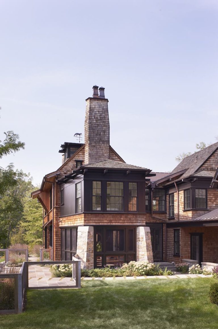 A large, brown Modern Shingle wooden house with black trim and a tall stone chimney. Multiple levels and big windows overlook a lush green lawn with stone walkways and landscaped flowerbeds. The sky above Princeton is clear and light blue.