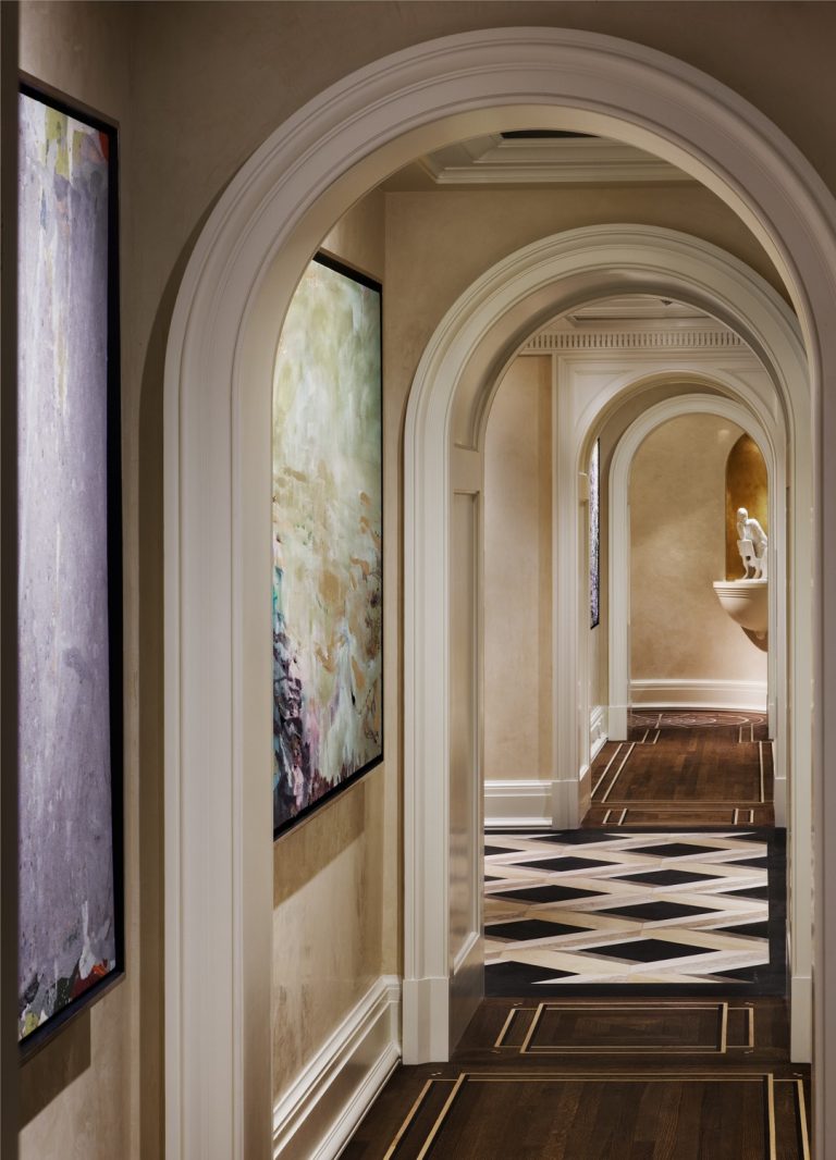 A hallway with cream-colored walls features several white arched doorways, modern abstract paintings on the walls, dark wood floors with light inlays, and black-and-white chevron tile in the distance, leading to a room with a white sculpture on a pedestal.