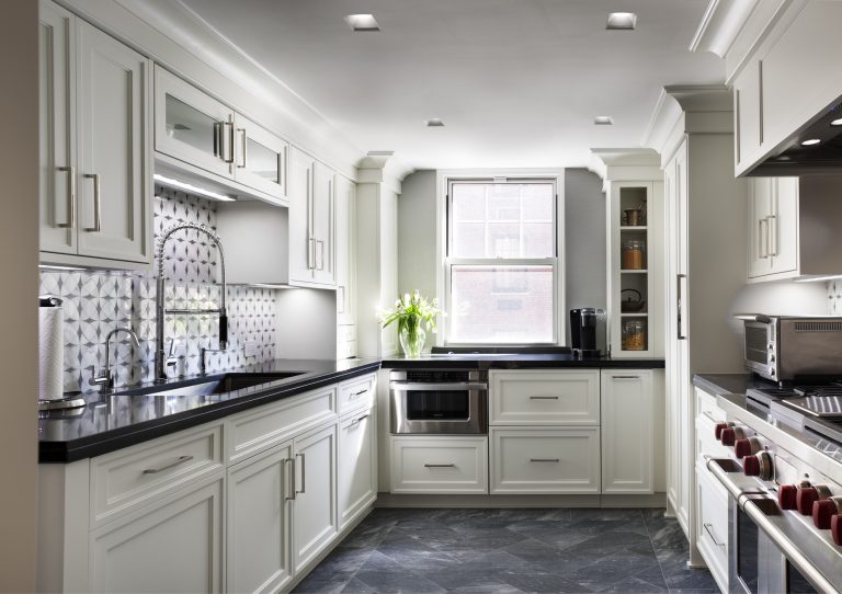 Bright, modern kitchen with white cabinets, black countertops, a patterned backsplash, stainless steel appliances, and a window letting in natural light. A vase of white flowers sits by the window, and the floor features dark gray tiles.