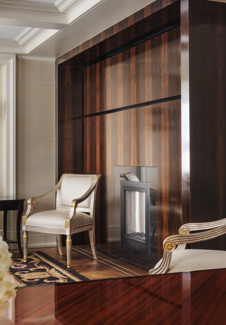 Elegant room with a dark wood-paneled fireplace, cream and gold upholstered armchairs, a patterned rug, and a polished wood table. The space feels luxurious with intricate molding on the ceiling and soft, neutral-toned walls.